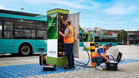 Techniker vor geöffneter Ladesäule und Busse im Hintergrund