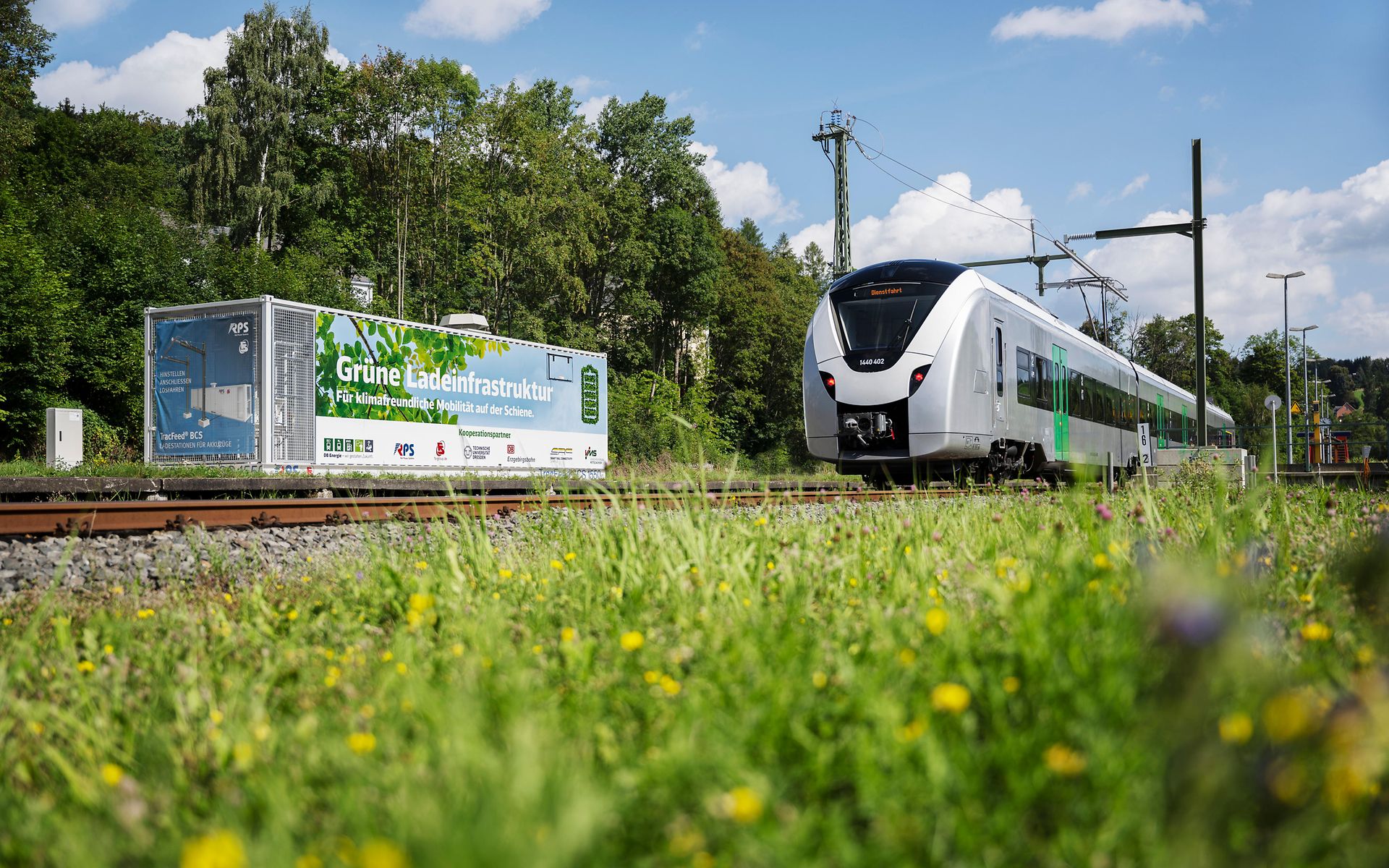 Ladeunterwerk neben Akkuzug (BEMU) in grüner Landschaft